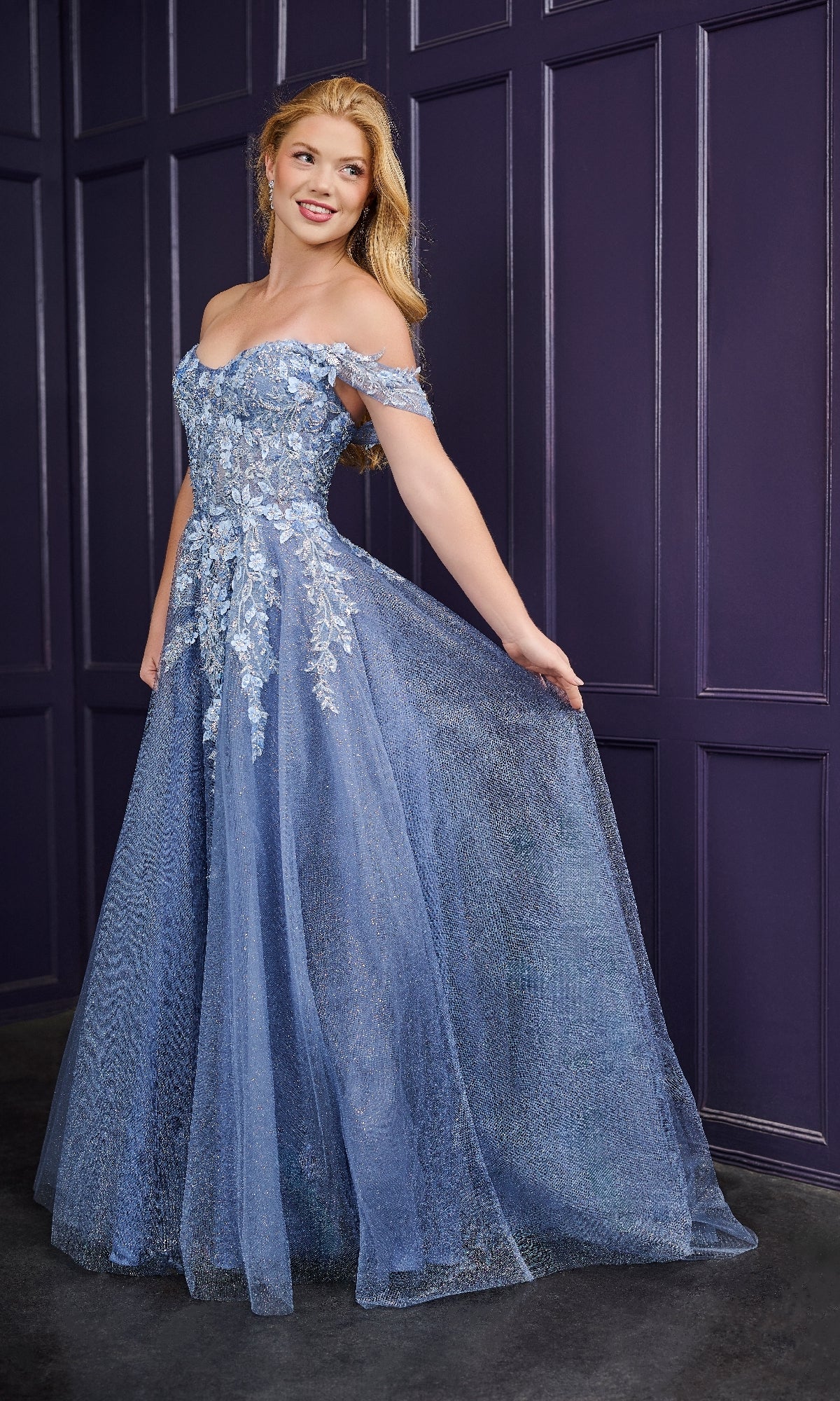 A woman models the Long Prom Dress: Tiffany 16243—an elegant blue off-the-shoulder gown with floral embroidery—as she poses gracefully against a purple paneled wall, holding out the skirt.